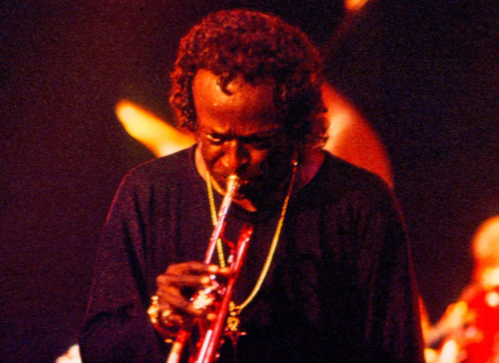 O trompetista Miles Davis tocando de cabeça baixa sob luz vermelha de palco