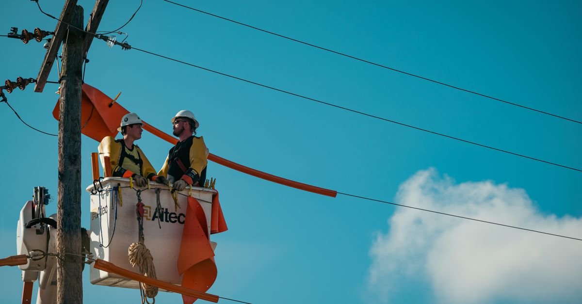 Dois eletricistas trabalham em cesto aéreo junto a poste de rede de energia elétrica sob céu azul