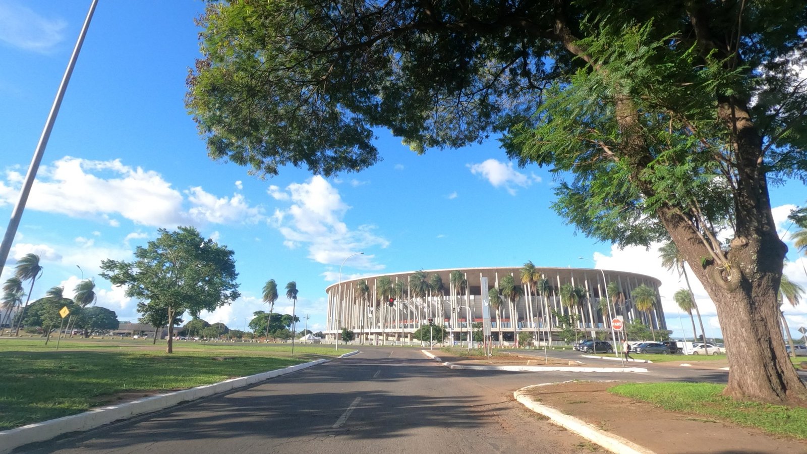 Fachada do Estádio Nacional Mané Garrincha vista de uma via do Eixo Monumental, com árvores e céu azul