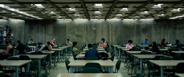 Cena do filme Mapas em salão amplo de concreto com mesas de estudo ocupadas