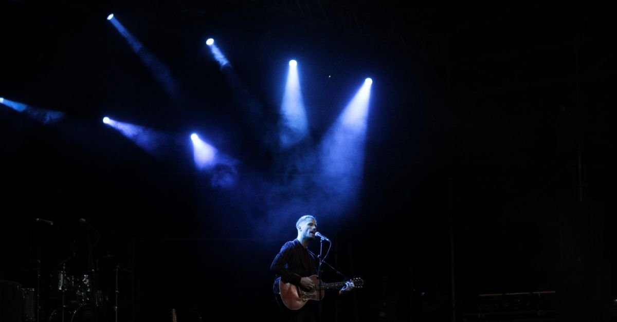 Músico toca violão e canta sozinho em palco iluminado por luz azul