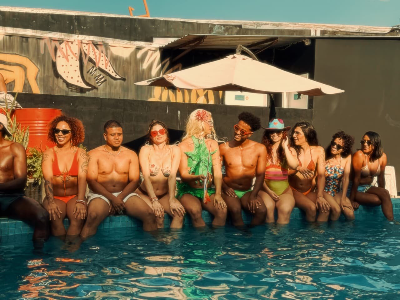 Grupo de pessoas sentado na borda de uma piscina em dia de sol, durante festa ao ar livre