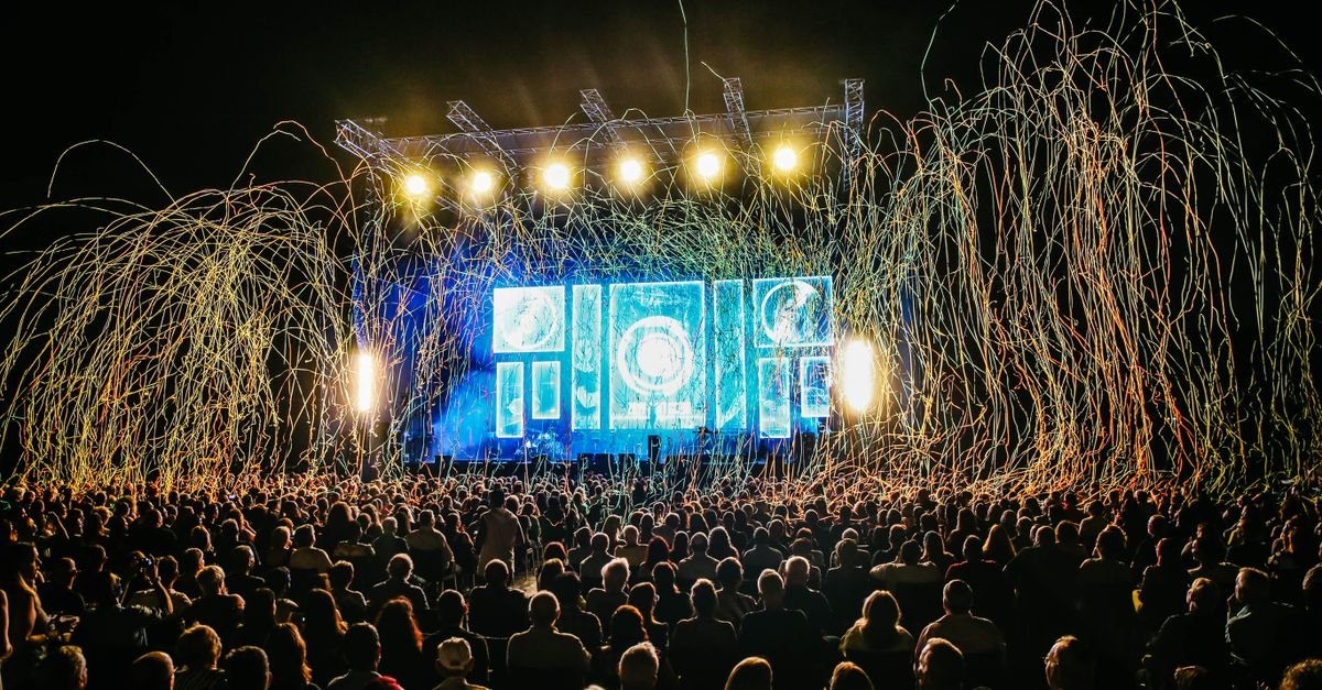 Público em festival de música à noite, com palco iluminado ao fundo
