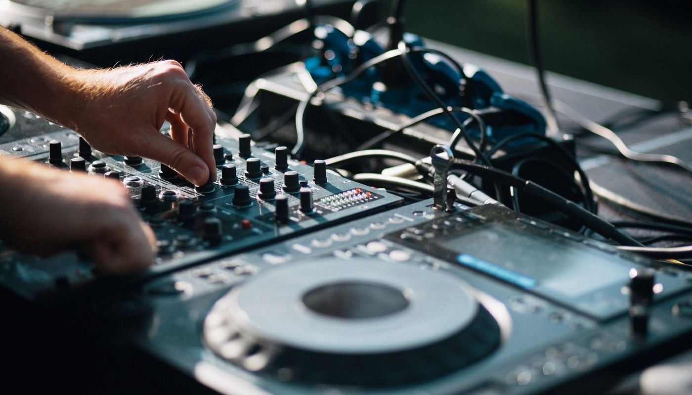 Mãos de um DJ ajustando os controles de uma mesa de mixagem ao lado de um CDJ durante apresentação ao ar livre