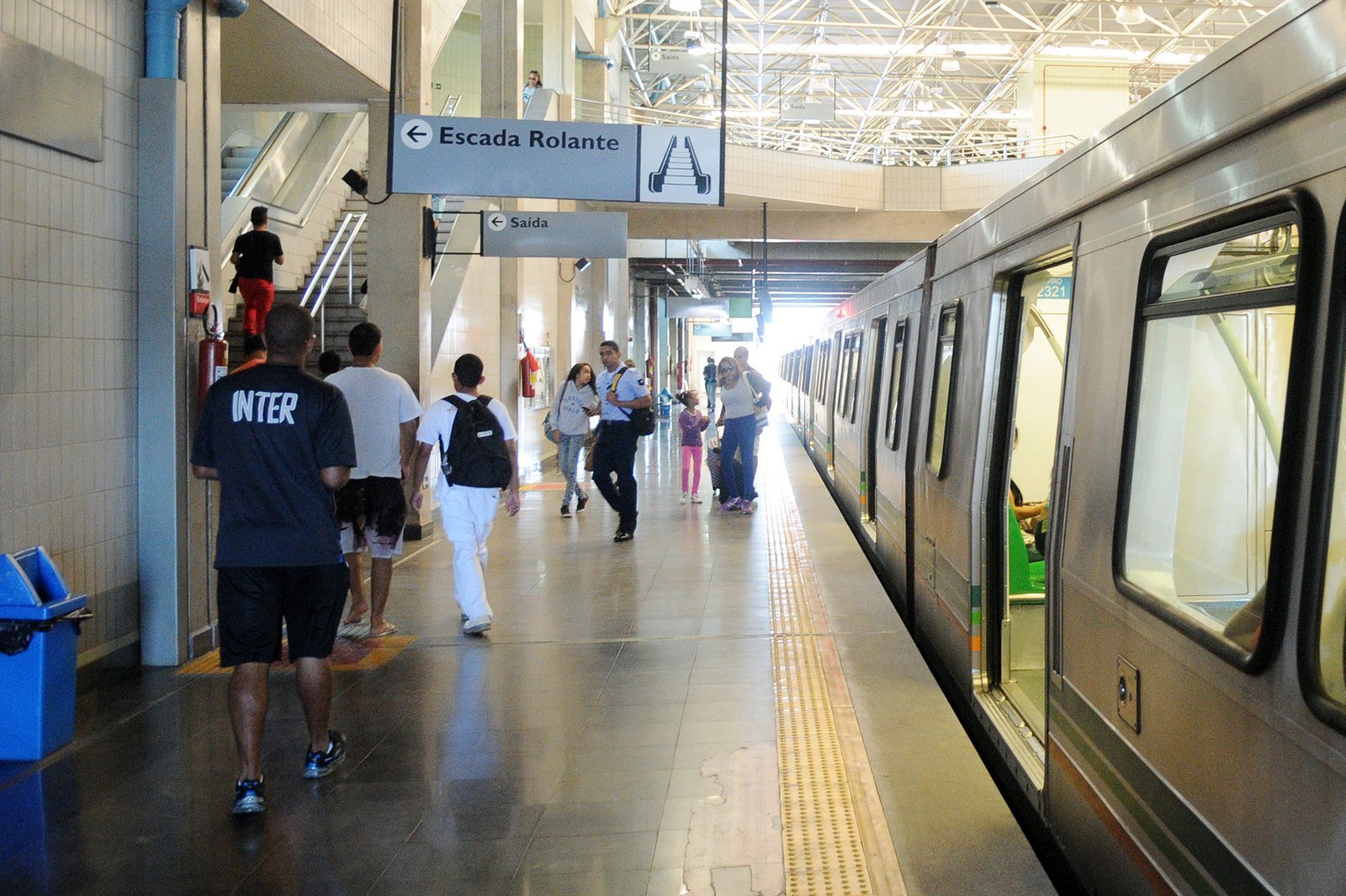 Plataforma do Metrô-DF com trem parado, placas de escada rolante e saída e passageiros caminhando
