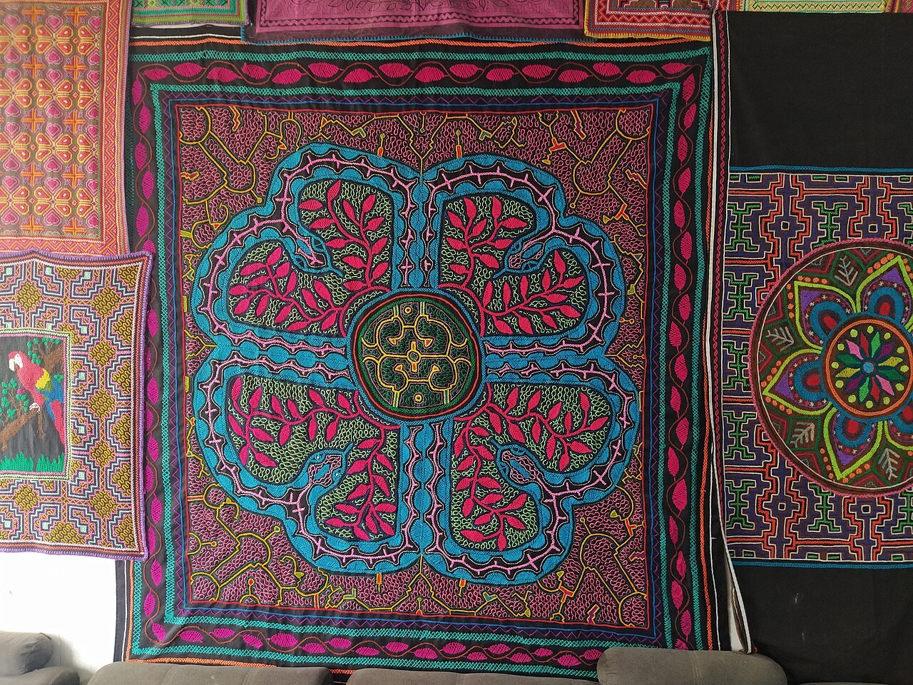Bordado Shipibo com padrão kené em azul, rosa e verde, pendurado na parede