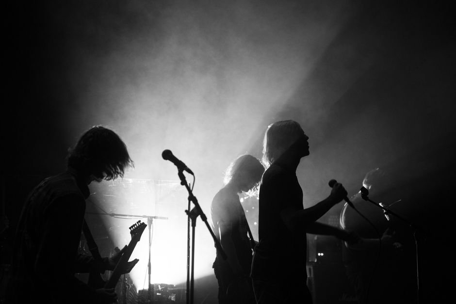 Silhuetas de músicos tocando ao vivo em palco com fumaça e contraluz, em foto preto e branco