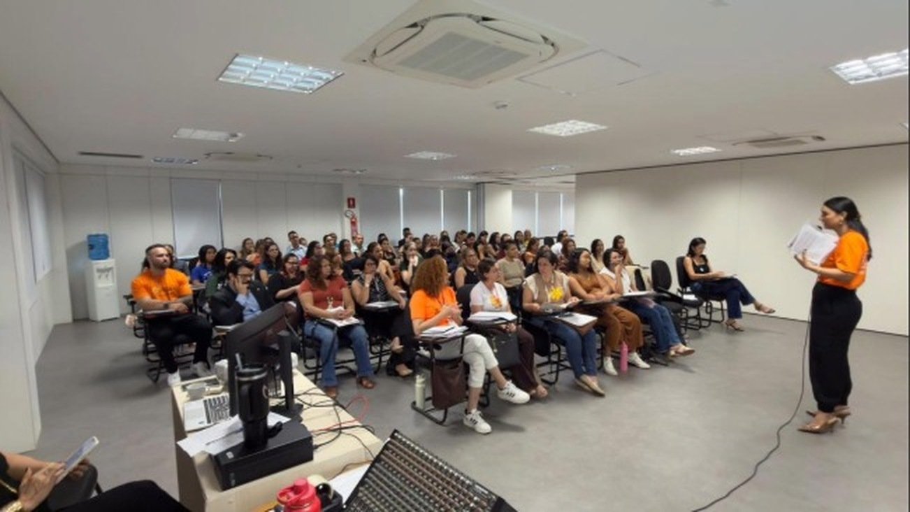 Sala com dezenas de educadores sentados assistindo a apresentação de palestrante em pé