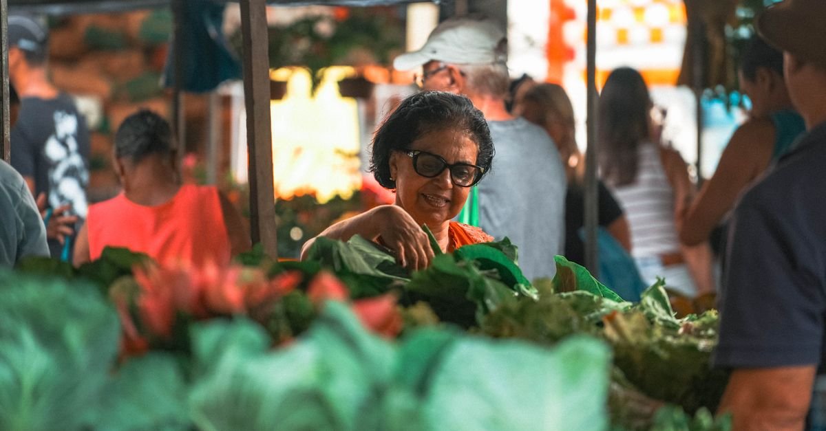 Mulher escolhe folhas verdes em banca de feira ao ar livre com verduras em primeiro plano
