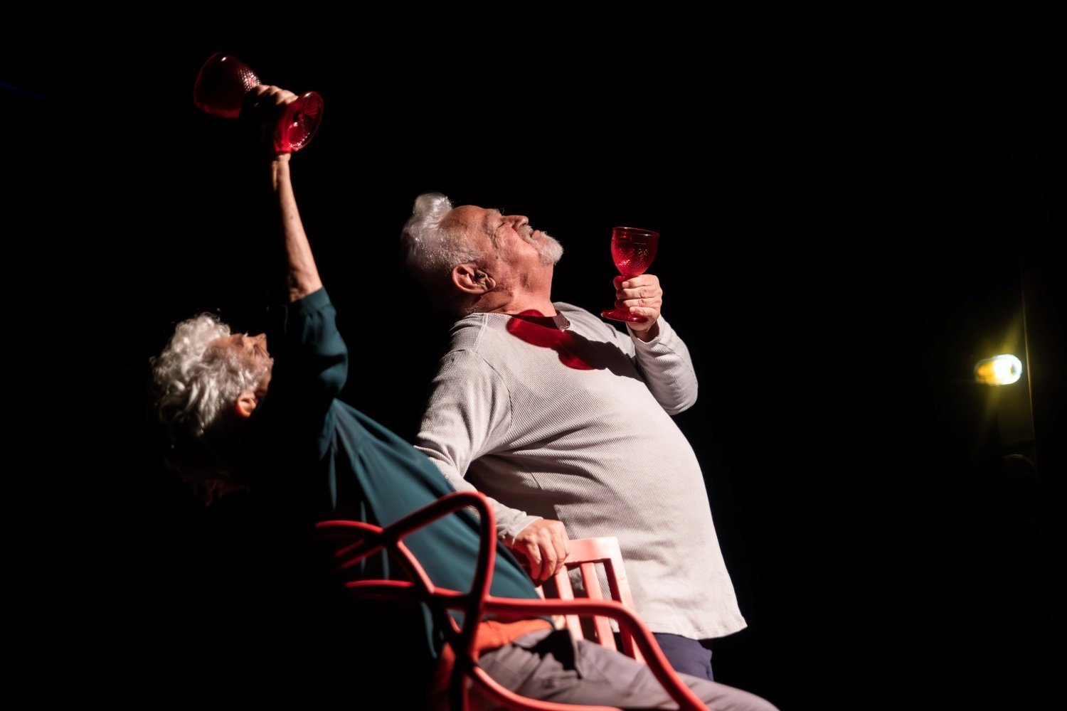 Dois atores idosos em cena, erguendo taças vermelhas sob luz de palco
