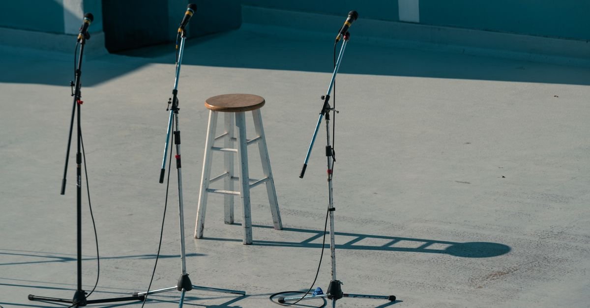 Palco com três pedestais de microfone e uma banqueta de madeira ao centro, sem público
