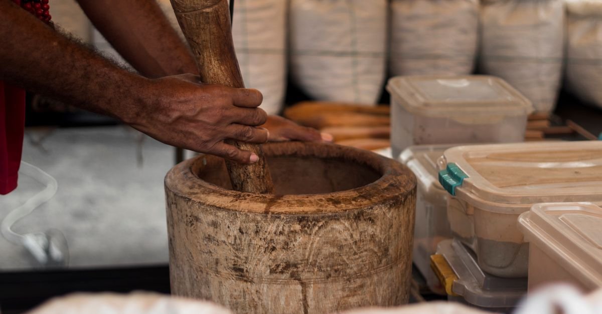 Mãos socam ingredientes em pilão de madeira em banca de feira de produtores