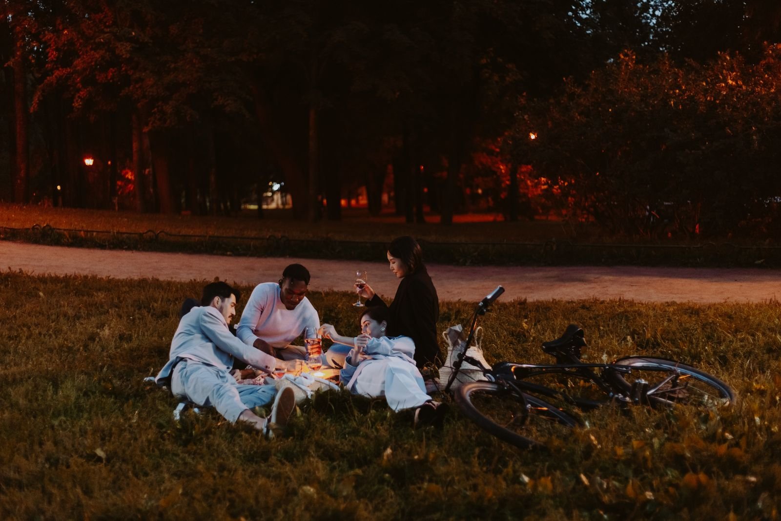 Amigos fazem piquenique em gramado de parque ao anoitecer, com uma bicicleta ao lado (foto ilustrativa)