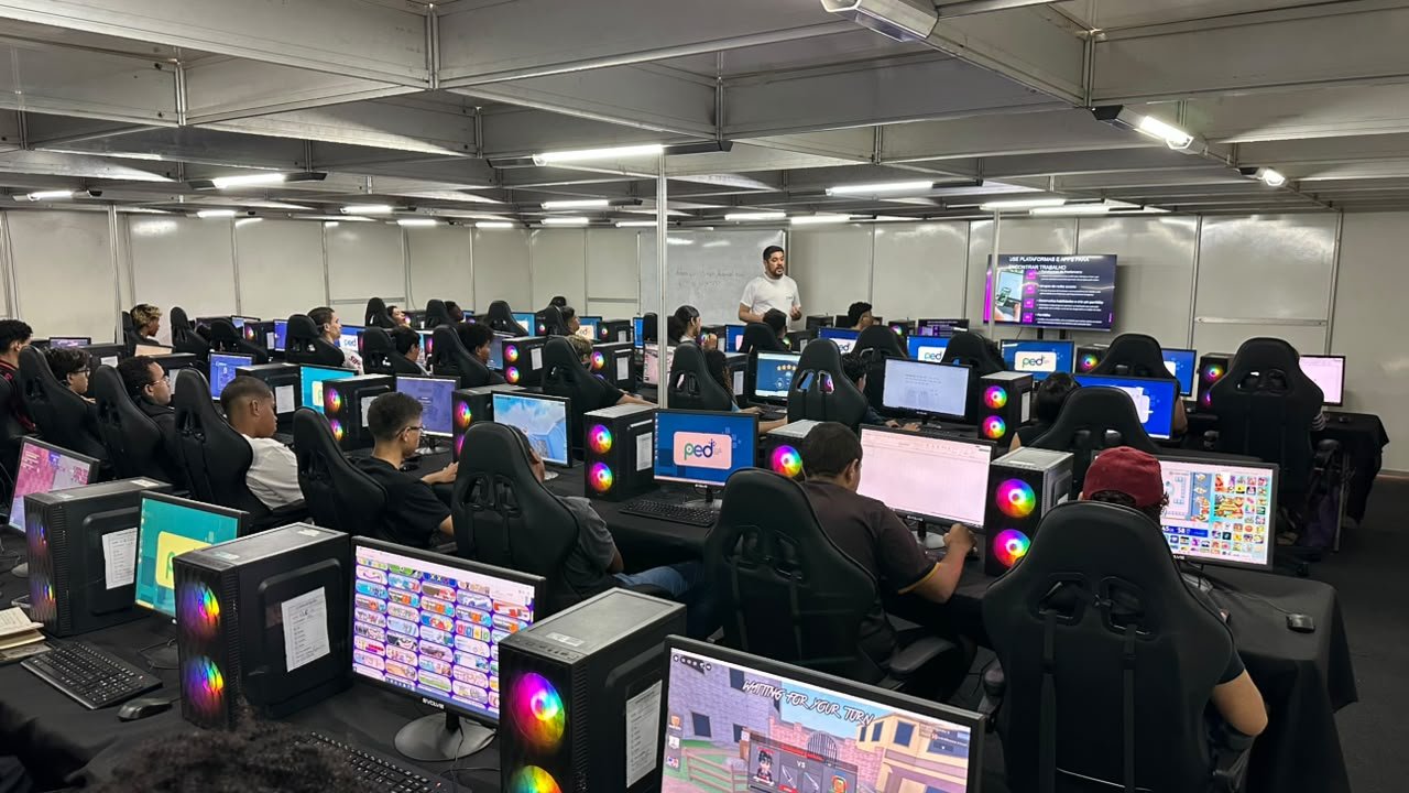 Instrutor em pé diante de uma sala com dezenas de alunos sentados em cadeiras gamer, cada um diante de um computador