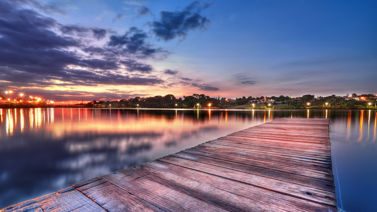 Píer de madeira sobre o Lago Paranoá ao entardecer, com as luzes da orla refletidas na água