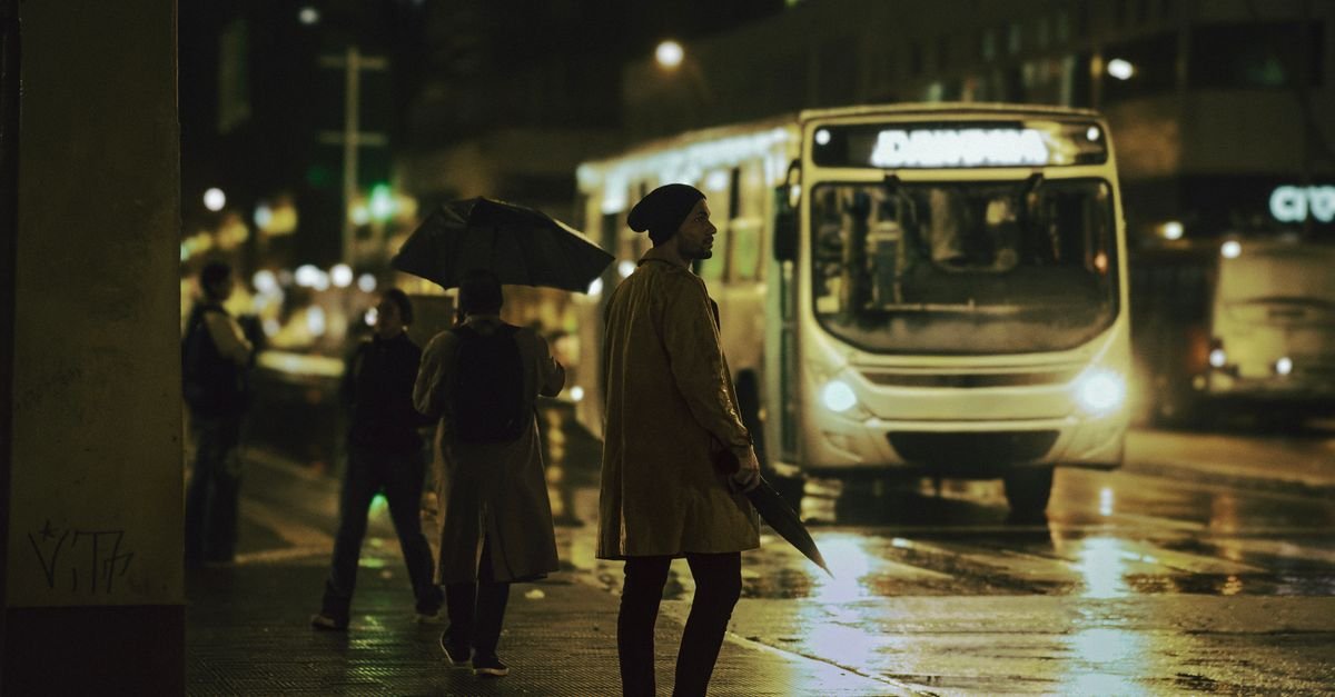 Ônibus urbano circula em avenida molhada à noite, com pedestres na calçada