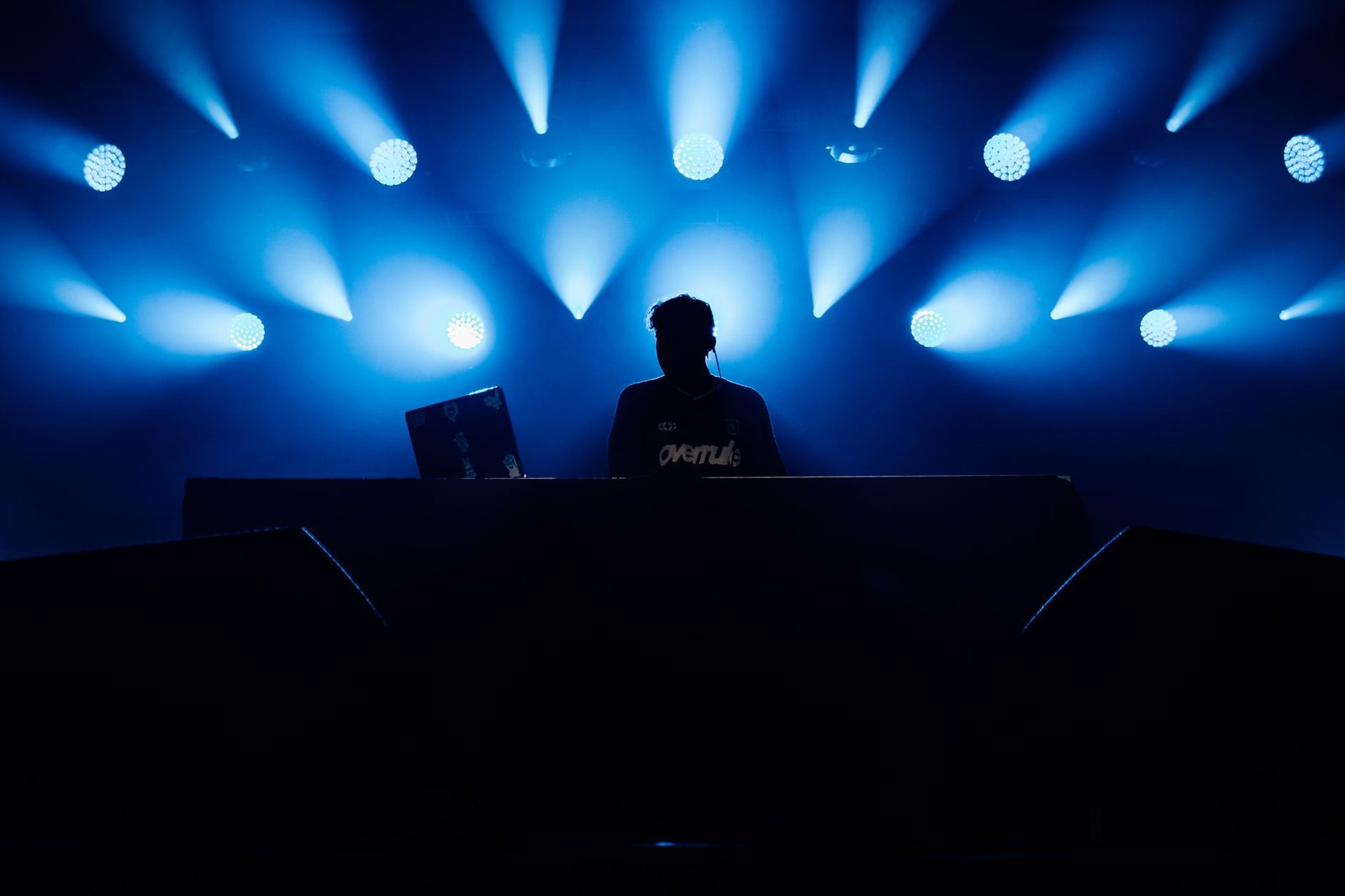 Silhueta de DJ atrás da mesa de som, diante de refletores azuis em palco de show