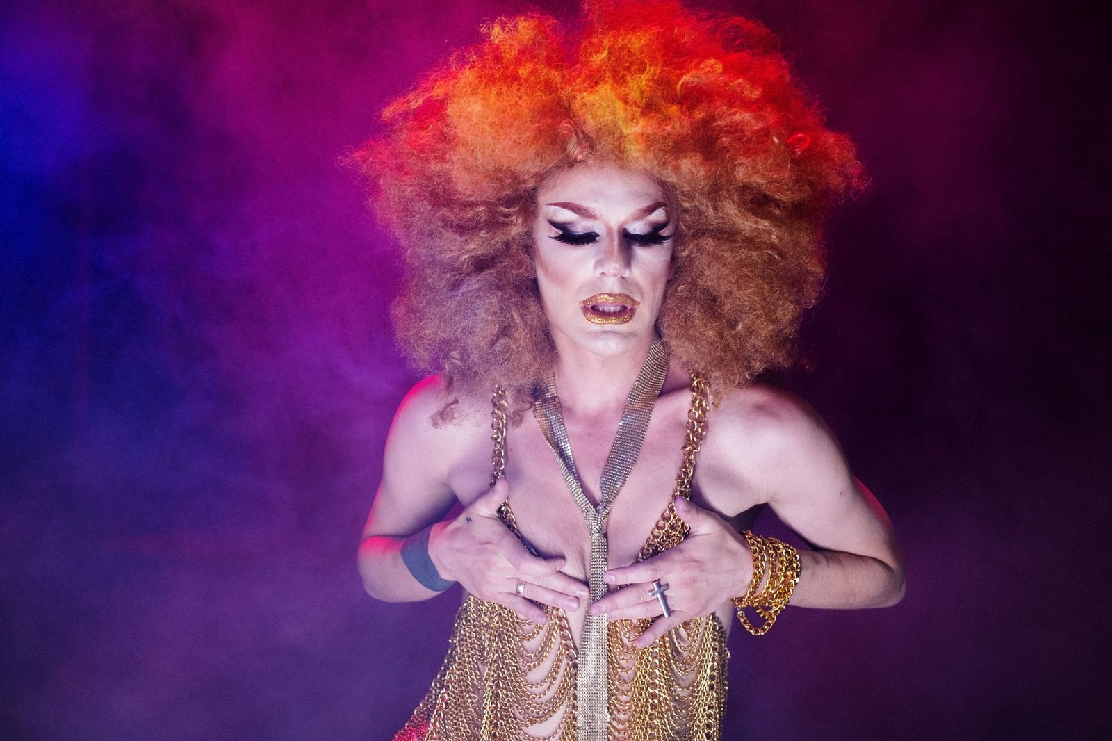 Artista drag de peruca volumosa laranja e figurino dourado sob luz roxa