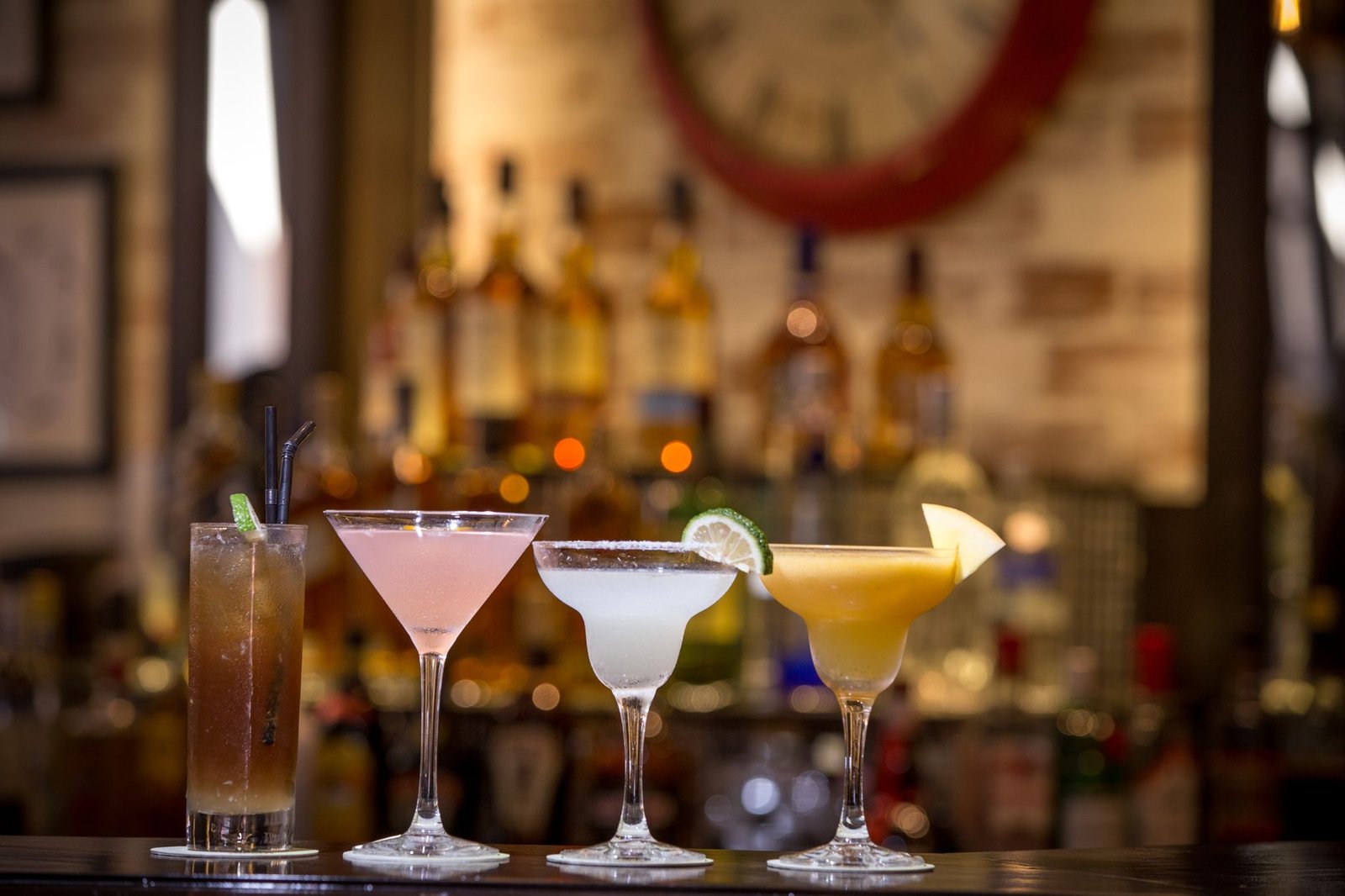Quatro coquetéis diferentes alinhados no balcão de um bar, com garrafas ao fundo