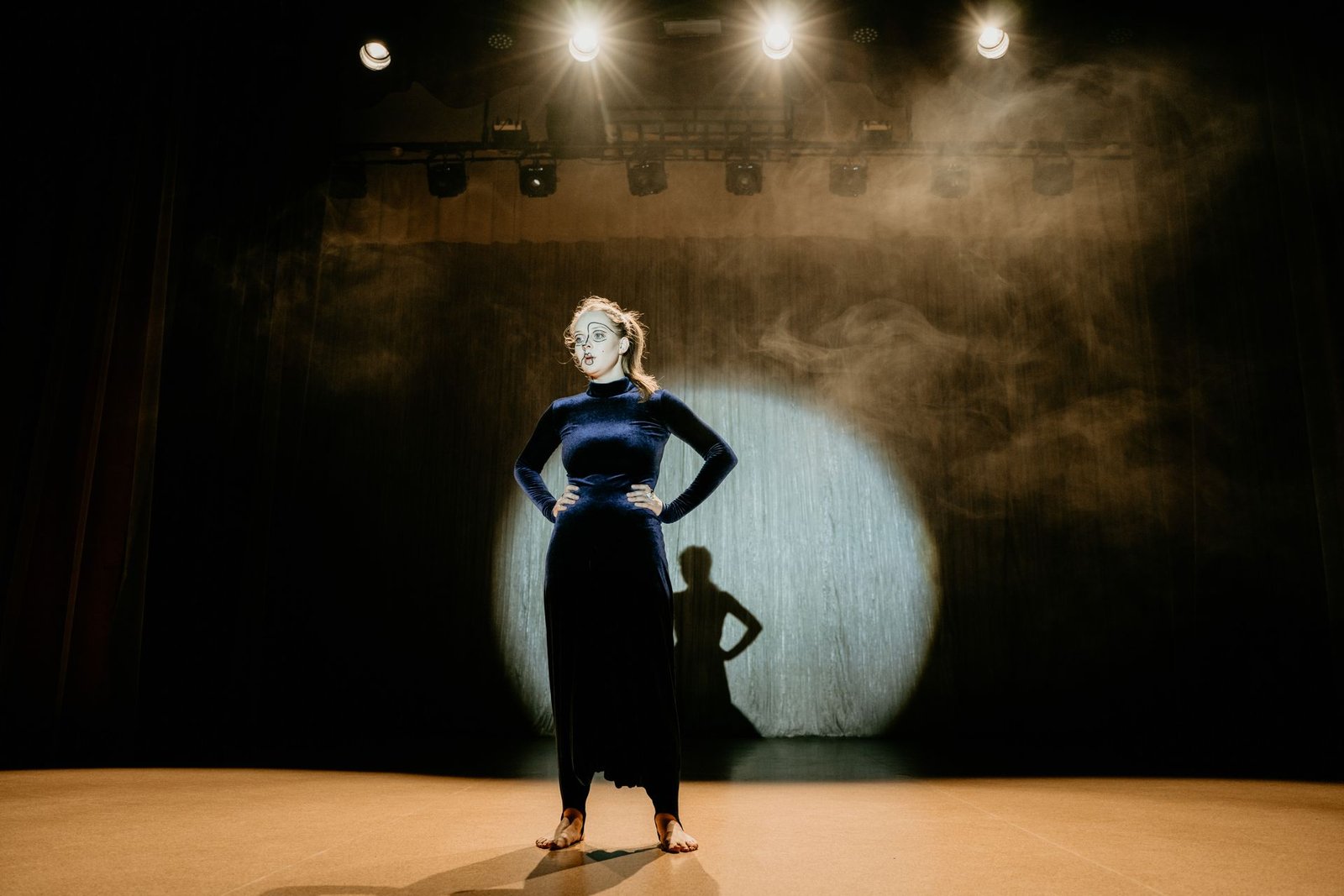 Atriz de maquiagem teatral posa de braços na cintura sob um facho de luz em um palco escuro, em foto ilustrativa de arquivo