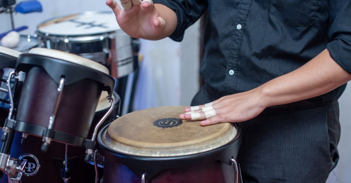 Mãos de músico tocando congas em ensaio, com bateria ao fundo