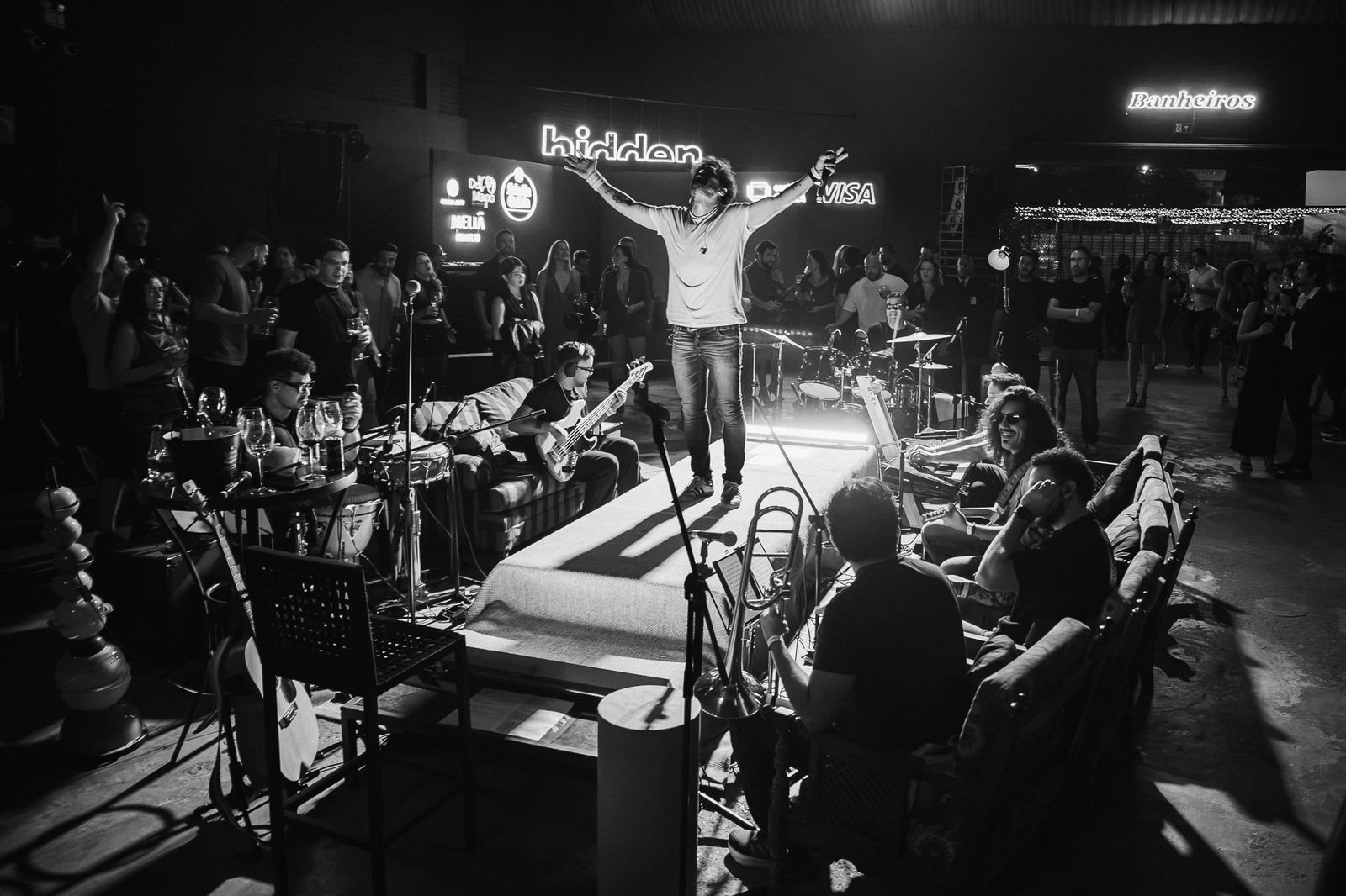 Cantor de braços abertos sobre palco baixo cercado pelo público, sob letreiro luminoso do Hidden Brasília