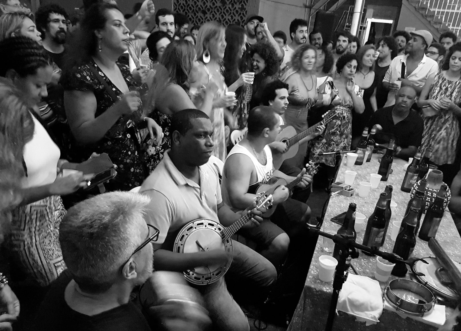 Roda de samba lotada, com músicos de cavaquinho e banjo sentados à mesa e público em pé ao redor, em foto de arquivo em preto e branco