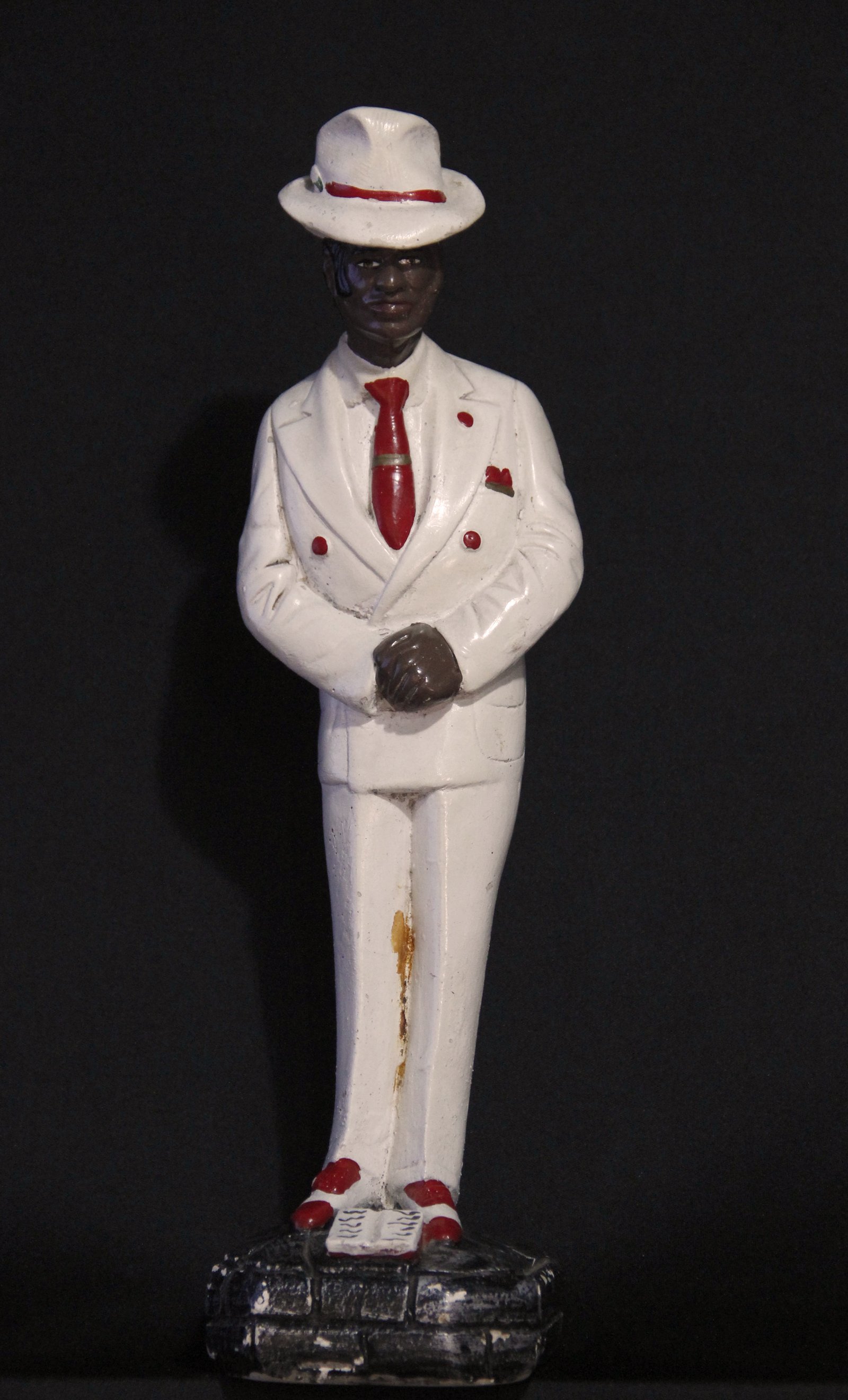Escultura de Zé Pelintra de terno branco, gravata vermelha e chapéu panamá, sobre fundo preto