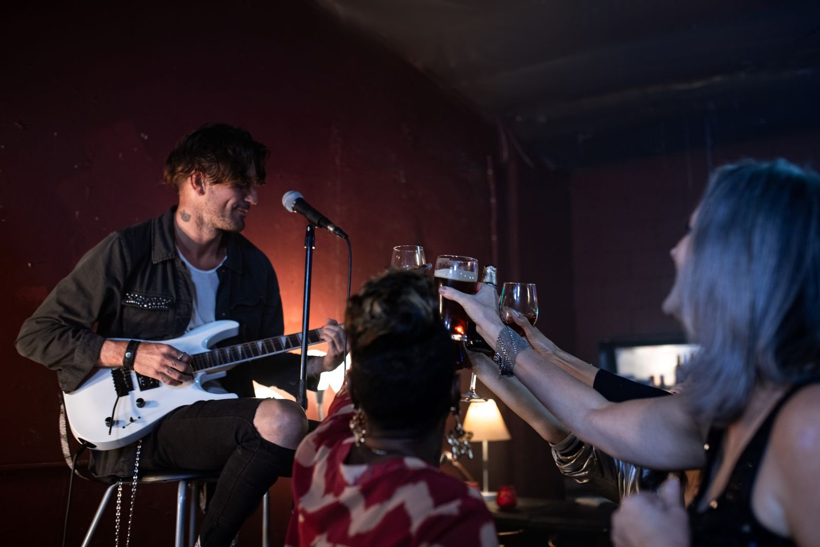 Músico toca em um palco pequeno de bar enquanto o público ergue copos num brinde (foto ilustrativa de banco de imagens)