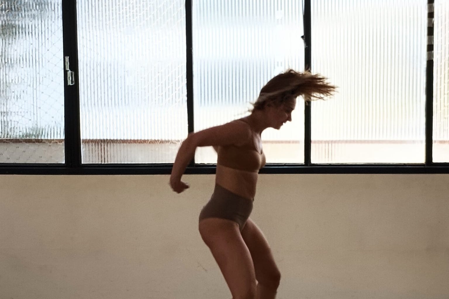 Bailarina corre de perfil em sala de ensaio iluminada por janelas, com o cabelo em movimento