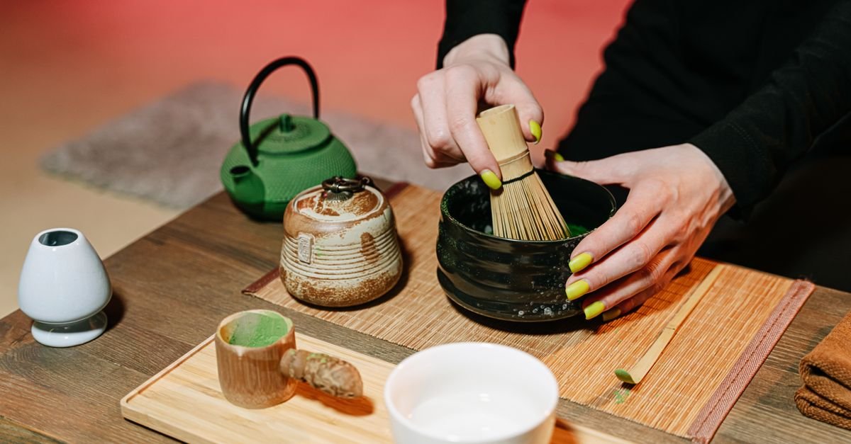 Mãos preparando chá matcha com batedor de bambu ao lado de bule de ferro verde e utensílios de cerâmica