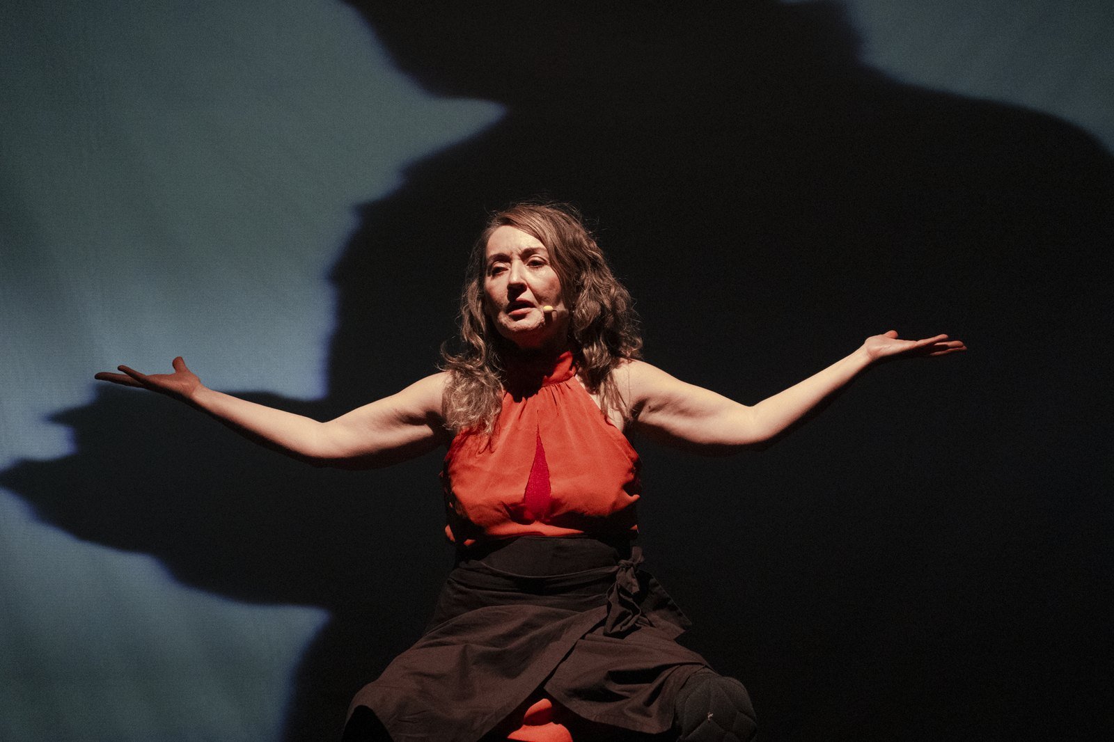 Atriz de blusa laranja abre os braços no palco, com a própria sombra projetada na parede ao fundo