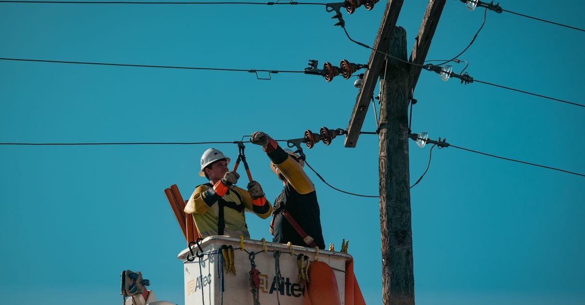 Dois eletricistas em cesto aéreo fazem manutenção em cabos de um poste de energia sob céu azul