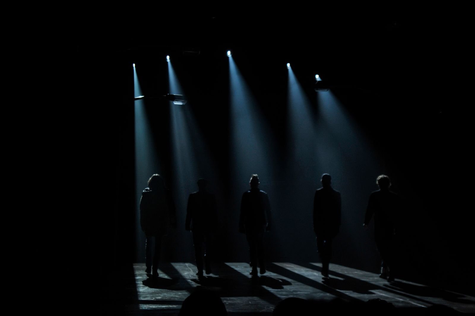Cinco silhuetas caminham num palco escuro sob feixes de luz, imagem de arquivo que ilustra teatro de sombras