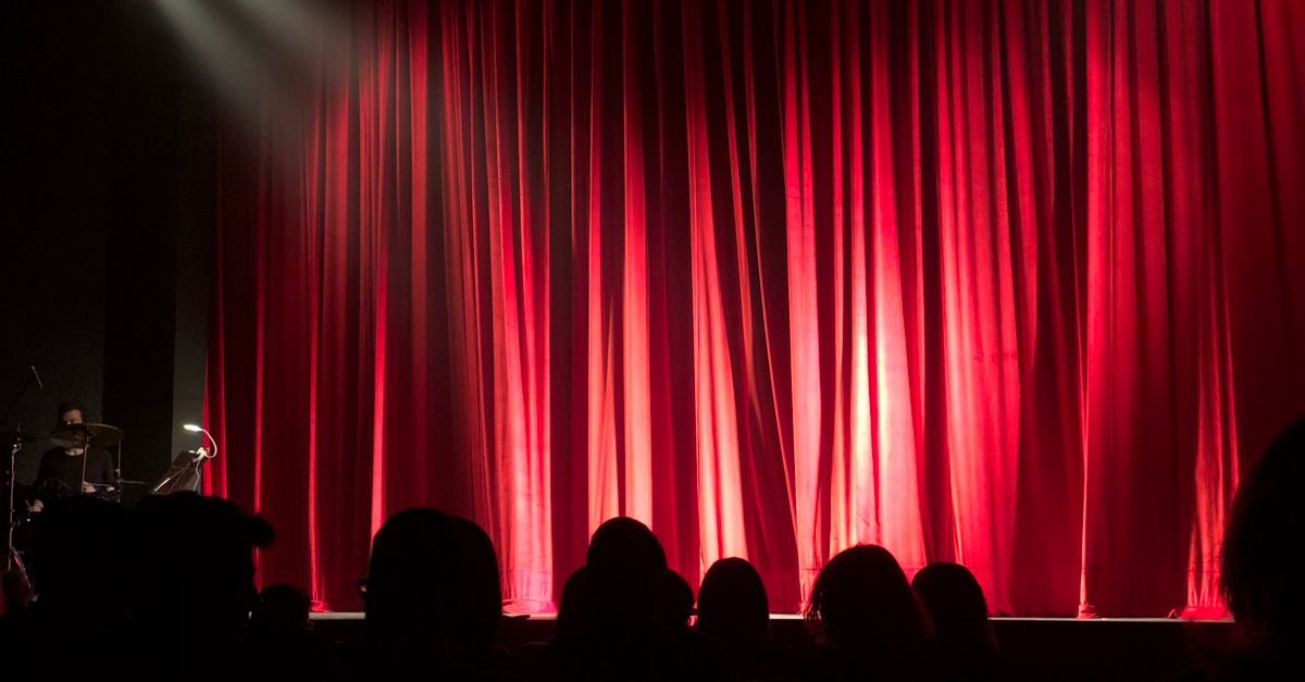 Palco de teatro com cortina vermelha iluminada e plateia em silhueta