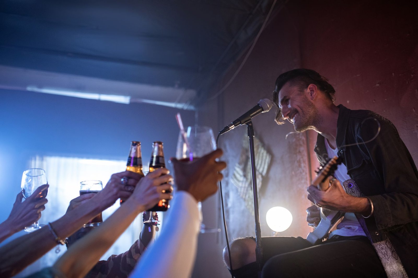 Cantor com violão ao microfone em palco de bar, com mãos brindando em primeiro plano