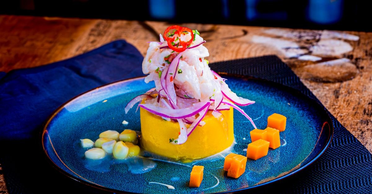 Ceviche peruano servido em prato azul, com cebola roxa, milho e cubos de batata-doce