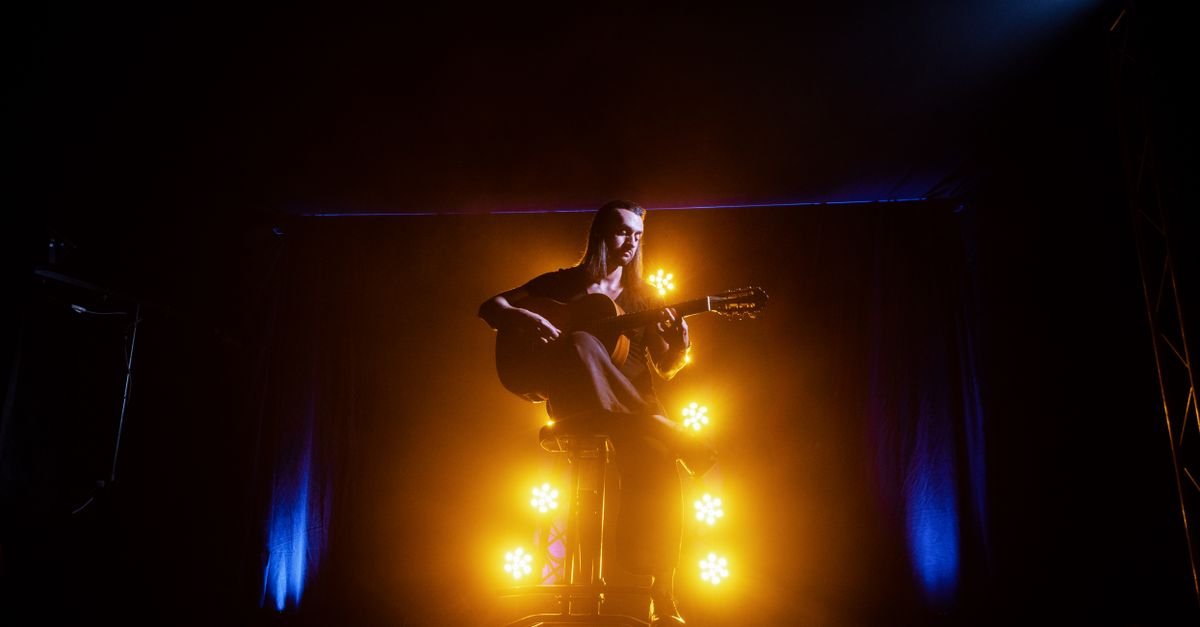 Músico toca violão sentado em um banco no palco, iluminado por luzes amarelas e azuis