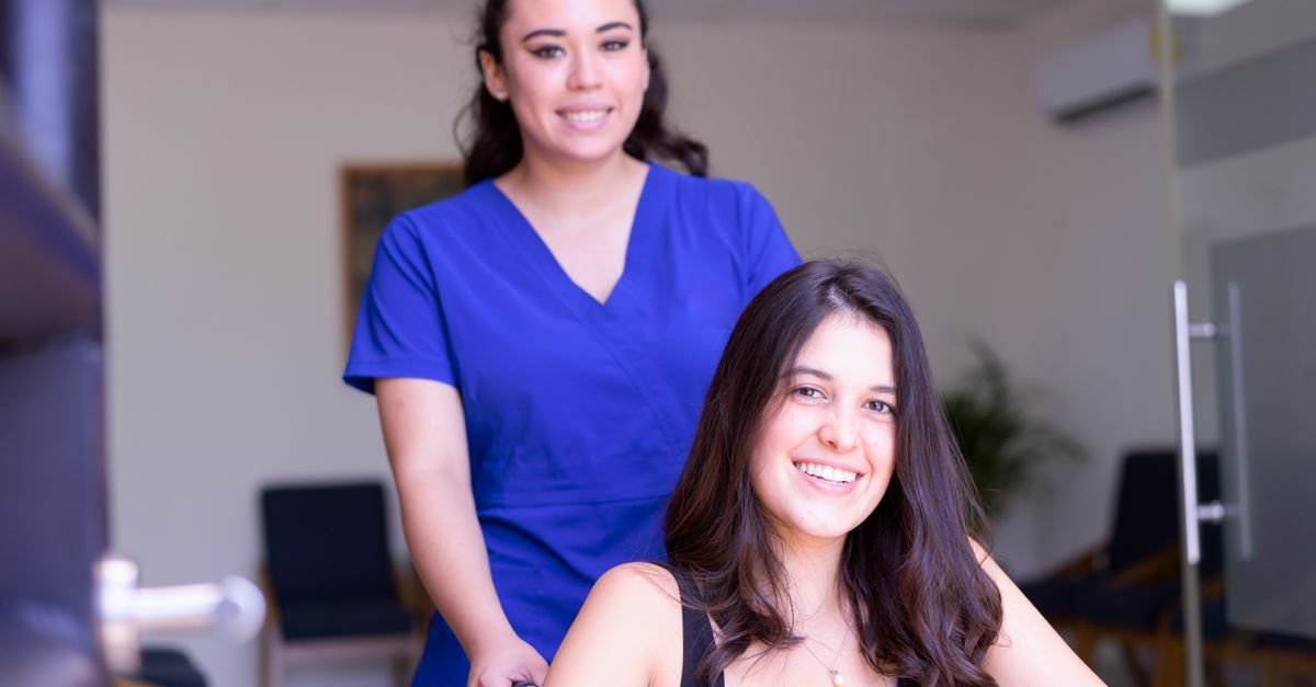 Enfermeira de uniforme azul conduz paciente em cadeira de rodas pelo corredor de uma unidade de saúde