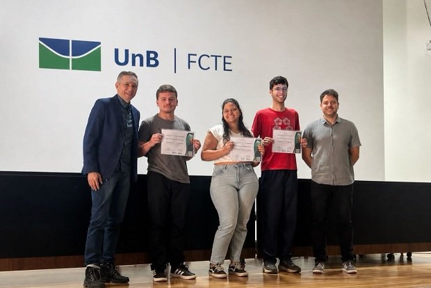 Três estudantes premiados seguram certificados ao lado de dois professores, diante do painel com a marca UnB FCTE