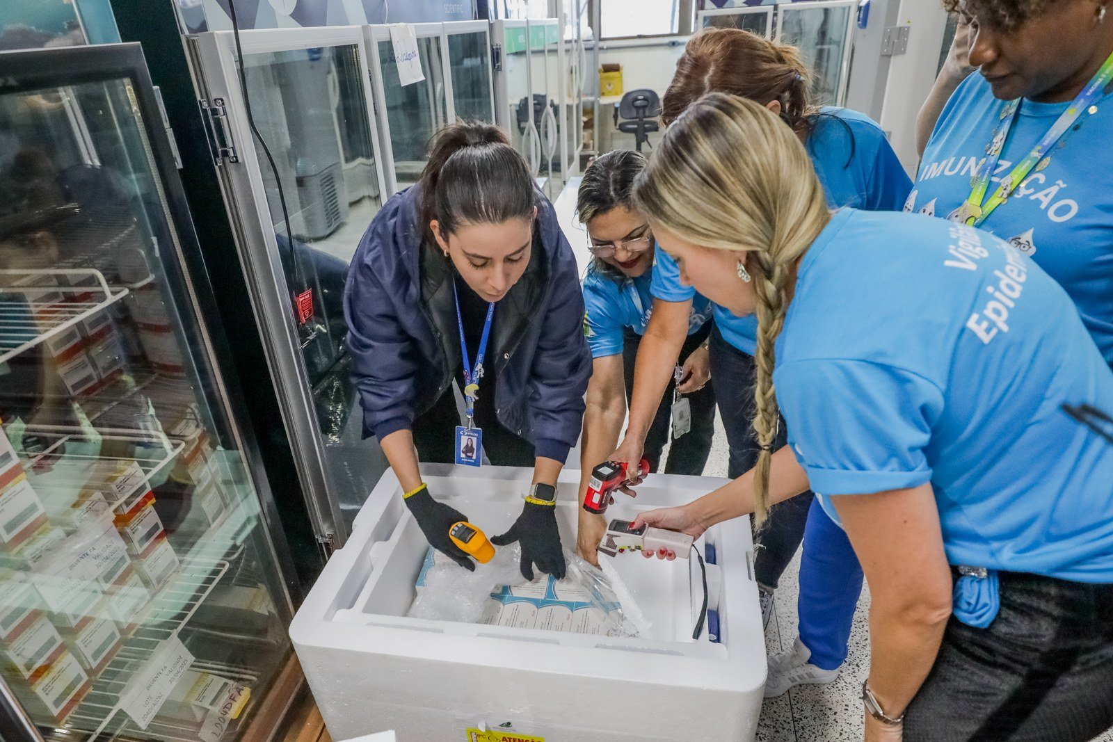 Equipe de vigilância epidemiológica confere a temperatura de doses em caixa térmica ao lado de geladeiras de vacinas