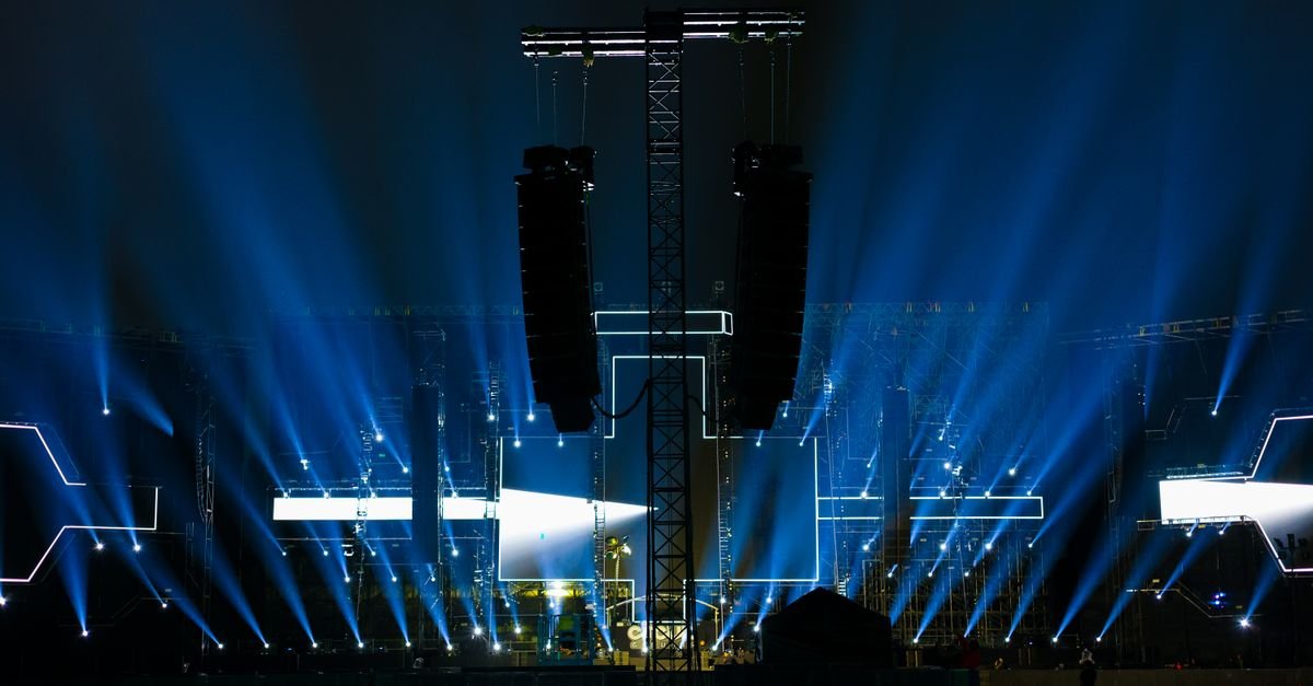 Palco de show ao ar livre a noite iluminado por feixes de luz azul e caixas de som suspensas