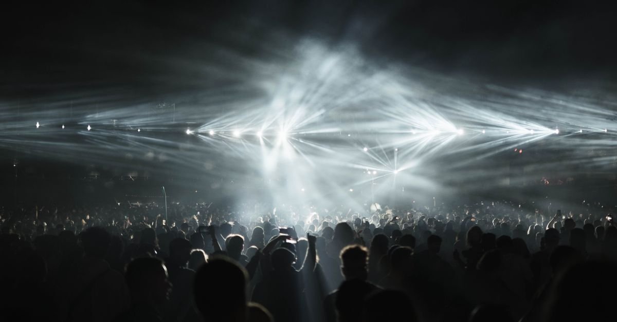 Público em silhueta diante de palco iluminado por feixes de luz em show noturno de música eletrônica