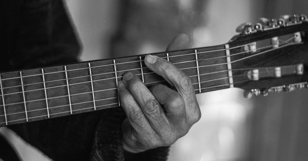 Mão de músico formando acorde no braço de um violão, em preto e branco