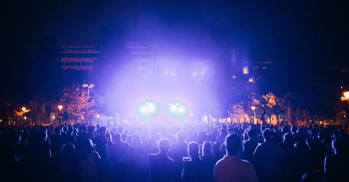 Público reunido diante de um palco iluminado por luzes roxas durante show ao ar livre à noite