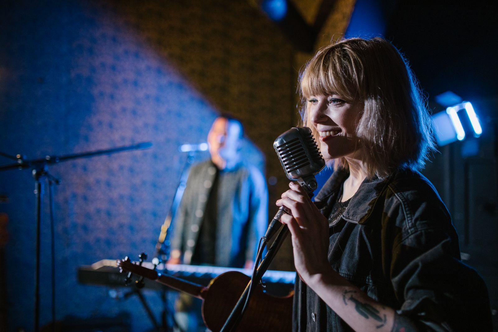 Cantora sorri diante de um microfone antigo em palco com luz azul, foto ilustrativa de show ao vivo